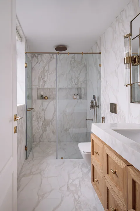 A narrow Mumbai apartment bathroom with a timber vanity, white marble surfaces, a fitted mirror and brass taps
